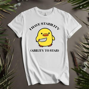 Pode incluir: Camiseta branca com um pintinho amarelo de desenho animado segurando uma faca. O texto acima do pintinho diz "I HAVE STABILITY" e abaixo, entre parênteses, "(ABILITY TO STAB)". Uma roupa casual.