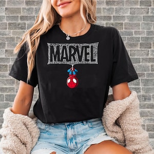 May include: Black t-shirt featuring the Marvel logo in white, with a spiderweb design. Below the logo, a cartoon Spider-Man figure hangs upside down. The shirt is a casual wear item.
