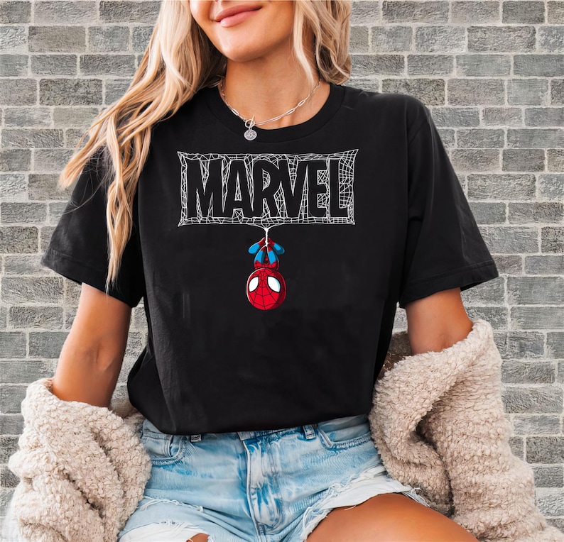 May include: Black t-shirt featuring the Marvel logo in white, with a spiderweb design. Below the logo, a cartoon Spider-Man figure hangs upside down. The shirt is a casual wear item.
