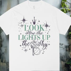 May include: White t-shirt with the text "LOOK How She LIGHTS UP the Sky" in green and purple. The design includes stars and a crown. The shirt is displayed against a rustic wooden and brick background.
