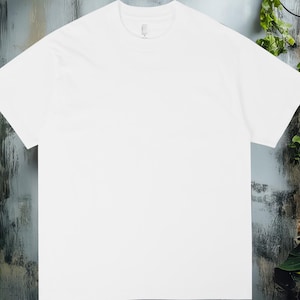 K&ouml;nnte beinhalten: Ein schlichtes wei&szlig;es T-Shirt mit Rundhalsausschnitt, vor dem Hintergrund einer verwitterten Wand und Gr&uuml;npflanzen. Das Shirt ist zentriert, um sein schlichtes Design und seine klaren Linien hervorzuheben. Der Hintergrund umfasst eine Backsteinmauer und &uuml;ppige gr&uuml;ne Pflanzen.