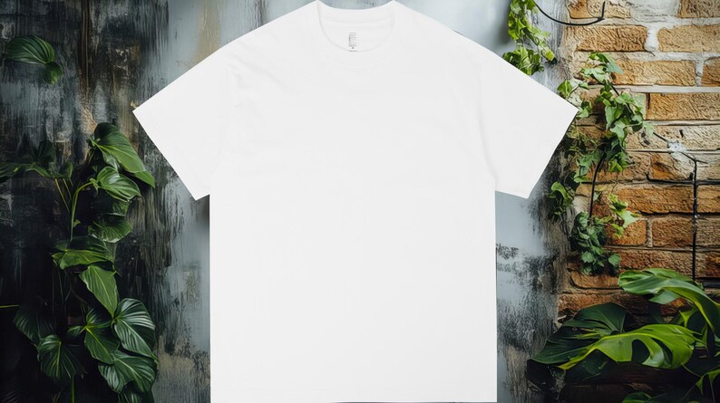 K&ouml;nnte beinhalten: Ein schlichtes wei&szlig;es T-Shirt mit Rundhalsausschnitt, vor dem Hintergrund einer verwitterten Wand und Gr&uuml;npflanzen. Das Shirt ist zentriert, um sein schlichtes Design und seine klaren Linien hervorzuheben. Der Hintergrund umfasst eine Backsteinmauer und &uuml;ppige gr&uuml;ne Pflanzen.
