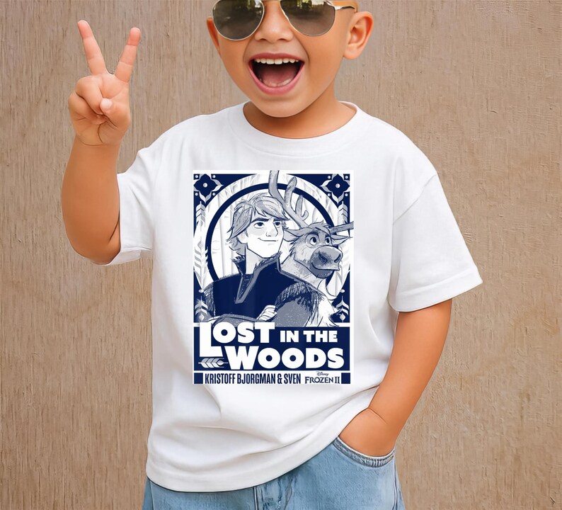 Puede incluir: Camiseta blanca con un gr&aacute;fico azul de Kristoff y Sven de Frozen II, con el texto "LOST IN THE WOODS". La camiseta es de cuello redondo y la lleva un ni&ntilde;o.