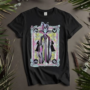 May include: Black t-shirt with a colorful illustration of Maleficent from Disney's Sleeping Beauty. The illustration is framed in a decorative border with floral accents. The text "Maleficent" is printed below the illustration.