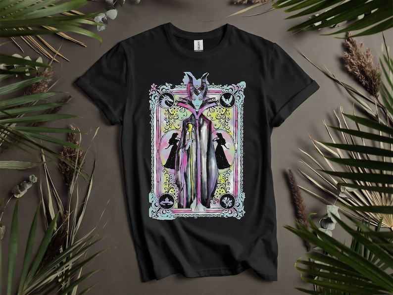 May include: Black t-shirt with a colorful illustration of Maleficent from Disney's Sleeping Beauty. The illustration is framed in a decorative border with floral accents. The text "Maleficent" is printed below the illustration.