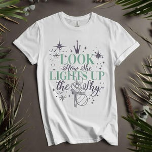 May include: White t-shirt with the text "Look How She Lights Up the Sky" in teal and purple. The design includes stars, a crown, and a whimsical illustration. A casual, everyday tee.
