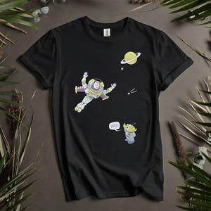 Może przedstawiać: Czarny t-shirt z kresk&oacute;wkową ilustracją Buzza Astrala w kosmosie, planetą i gwiazdami. Obecna jest r&oacute;wnież postać kosmity z napisem "oooo" w dymku. Koszulka otoczona jest zielonymi liśćmi.