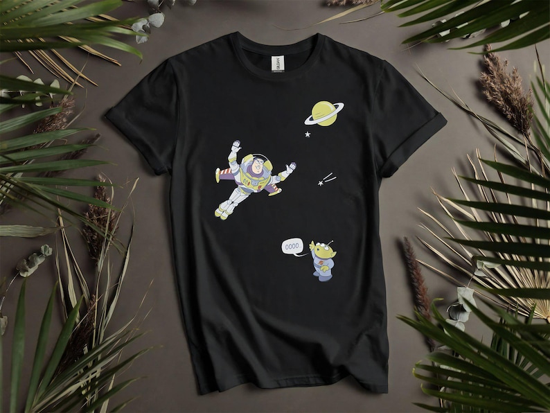 Może przedstawiać: Czarny t-shirt z kresk&oacute;wkową ilustracją Buzza Astrala w kosmosie, planetą i gwiazdami. Obecna jest r&oacute;wnież postać kosmity z napisem "oooo" w dymku. Koszulka otoczona jest zielonymi liśćmi.
