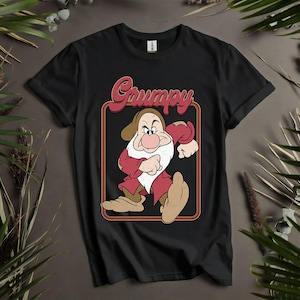 Puede incluir: Camiseta negra con una ilustración de dibujos animados de Gruñón de Blancanieves. El personaje lleva un atuendo rojo y marrón y tiene una barba blanca, dentro de un marco rectangular. La palabra "Grumpy" está escrita encima del personaje.