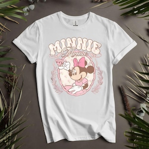 Puede incluir: Camiseta blanca con un dise&ntilde;o de dibujos animados de Minnie Mouse. El dise&ntilde;o muestra a Minnie Mouse sosteniendo una taza de t&eacute;, dentro de un marco ovalado rosa, con las palabras "Minnie Mouse" en una fuente retro. La camiseta est&aacute; rodeada de hojas verdes.