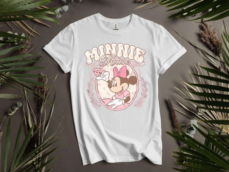 Puede incluir: Camiseta blanca con un dise&ntilde;o de dibujos animados de Minnie Mouse. El dise&ntilde;o muestra a Minnie Mouse sosteniendo una taza de t&eacute;, dentro de un marco ovalado rosa, con las palabras "Minnie Mouse" en una fuente retro. La camiseta est&aacute; rodeada de hojas verdes.