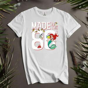 May include: White t-shirt with the text "MADE IN THE 80'S" in red and white. The number 8 is designed with a mermaid and other cartoon characters. The shirt is a short-sleeved crew neck.