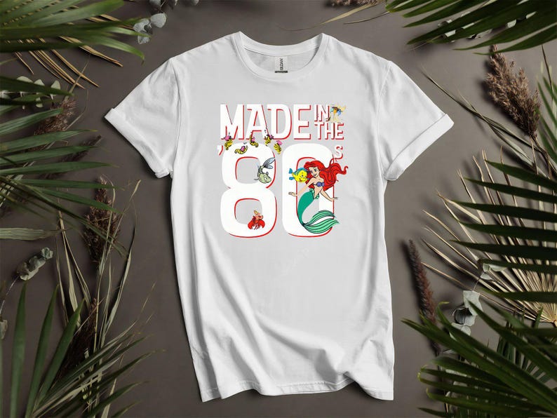 May include: White t-shirt with the text "MADE IN THE 80'S" in red and white. The number 8 is designed with a mermaid and other cartoon characters. The shirt is a short-sleeved crew neck.