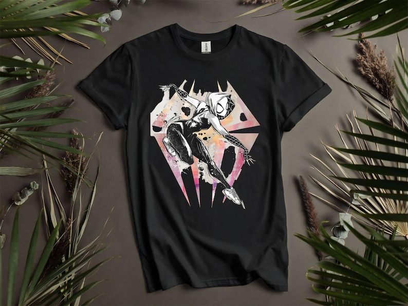 May include: Black t-shirt featuring a graphic of a superhero in a dynamic pose. The design is in black and white with watercolor-style accents of pink, orange, and yellow. The superhero is set against a geometric background.