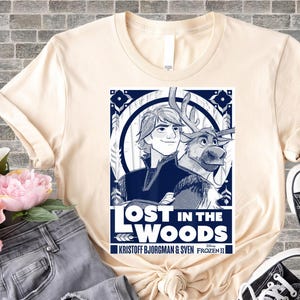 Puede incluir: Camiseta color crema con un gr&aacute;fico azul y blanco de la pel&iacute;cula Frozen II. El gr&aacute;fico incluye el texto "LOST IN THE WOODS" e im&aacute;genes de Kristoff, Sven y elementos decorativos. La camiseta se combina con vaqueros y zapatillas.