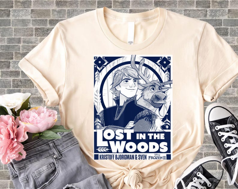 Puede incluir: Camiseta color crema con un gr&aacute;fico azul y blanco de la pel&iacute;cula Frozen II. El gr&aacute;fico incluye el texto "LOST IN THE WOODS" e im&aacute;genes de Kristoff, Sven y elementos decorativos. La camiseta se combina con vaqueros y zapatillas.