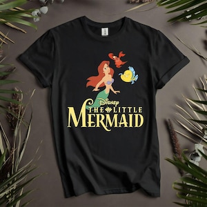 Puede incluir: Camiseta negra con un gr&aacute;fico colorido de Ariel, Flounder y Sebasti&aacute;n de La Sirenita de Disney. El t&iacute;tulo est&aacute; en escritura amarilla. La camiseta se muestra sobre un fondo neutro con vegetaci&oacute;n.