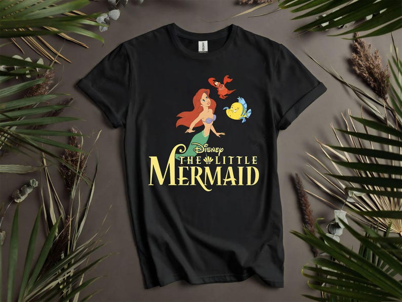Puede incluir: Camiseta negra con un gr&aacute;fico colorido de Ariel, Flounder y Sebasti&aacute;n de La Sirenita de Disney. El t&iacute;tulo est&aacute; en escritura amarilla. La camiseta se muestra sobre un fondo neutro con vegetaci&oacute;n.