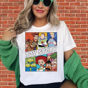 May include: White t-shirt featuring a colorful graphic of characters from the animated film "Toy Story." The design includes Woody, Buzz Lightyear, Bo Peep, and other characters, with the words "Toy Story" in bold letters.