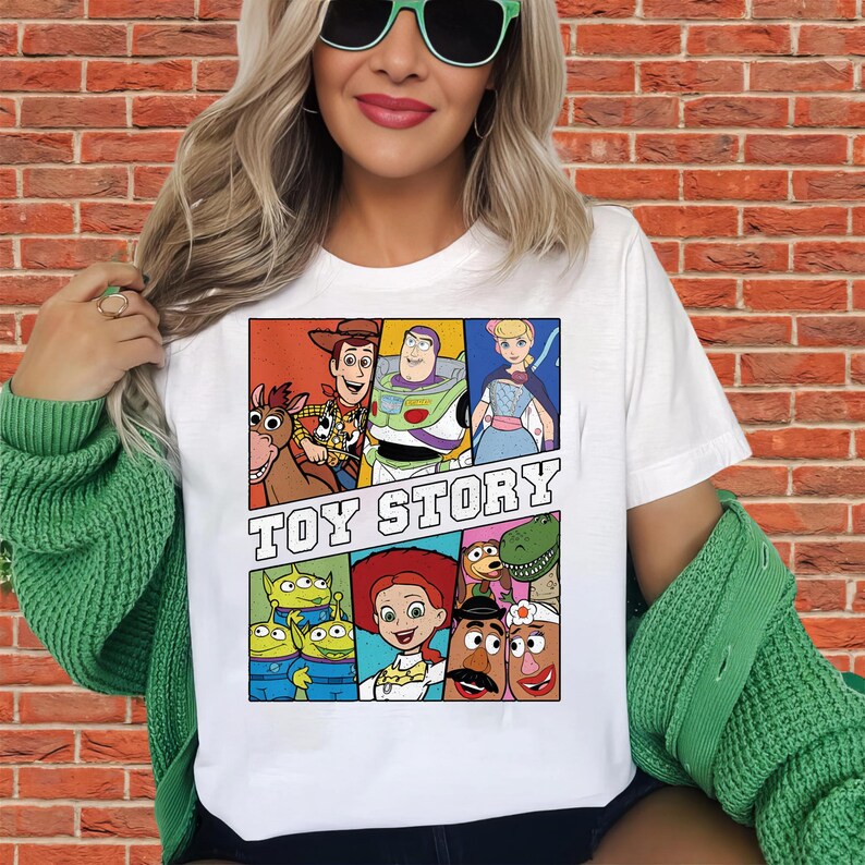 May include: White t-shirt featuring a colorful graphic of characters from the animated film "Toy Story." The design includes Woody, Buzz Lightyear, Bo Peep, and other characters, with the words "Toy Story" in bold letters.