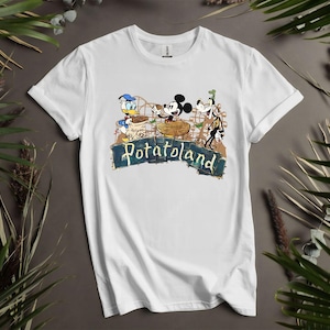May include: White t-shirt featuring a colorful graphic of Donald Duck, Mickey Mouse, and Goofy on a roller coaster. The design includes the text "Potatoland" in a distressed font, evoking a vintage amusement park theme.