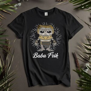May include: Black t-shirt featuring a cartoon illustration of a character from the Star Wars series, wearing a helmet and holding a tool. The text "Babu Frik" is printed below the illustration.