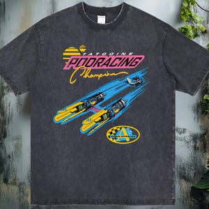 以下が含まれることがあります： レトロなグラフィックデザインのダークグレーのTシャツ。デザインは、ピンク、イエロー、ブルーで「TATOOINE PODRACING Champion」というテキストと、ポッドレーサーの画像が特徴です。このシャツはヴィンテージで使い古された外観をしています。