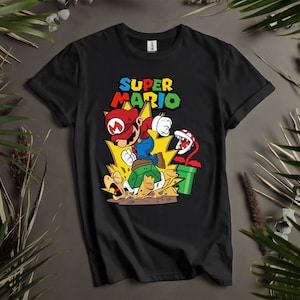 Puede incluir: Camiseta negra con un gráfico de Super Mario saltando sobre una tortuga. El gráfico también incluye una planta Piraña y una tubería verde. El texto "SUPER MARIO" está impreso en letras amarillas sobre el gráfico.