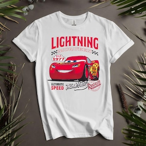 May include: White t-shirt featuring a graphic of Lightning McQueen from the movie Cars. The design includes the text "LIGHTNING McQUEEN" above a red race car, with "est. 1977" and "ULTIMATE SPEED Think Fast Rusterze" below.