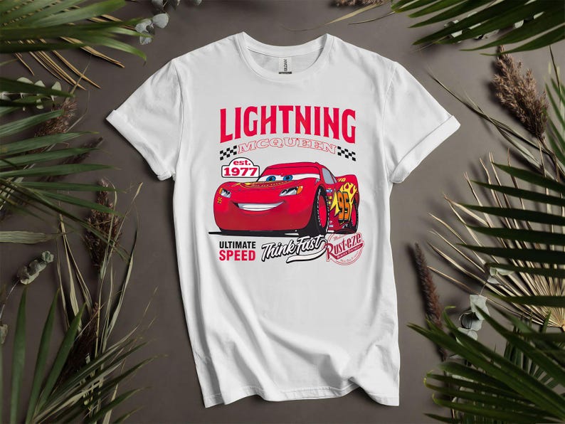 May include: White t-shirt featuring a graphic of Lightning McQueen from the movie Cars. The design includes the text "LIGHTNING McQUEEN" above a red race car, with "est. 1977" and "ULTIMATE SPEED Think Fast Rusterze" below.