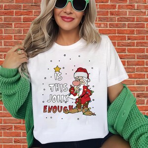 以下が含まれることがあります： 赤いスーツと帽子を着た漫画のクリスマスキャラクターが、クリスマスライトを持っている白いTシャツ。Tシャツには「IS THIS JOLLY ENOUGH？」の文字がプリントされています。Tシャツは人が着ています。