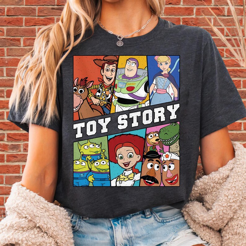 May include: Dark gray t-shirt featuring a colorful graphic of characters from the animated film "Toy Story." The design includes Woody, Buzz Lightyear, Bo Peep, and other beloved characters. The words "TOY STORY" are printed in bold white letters.