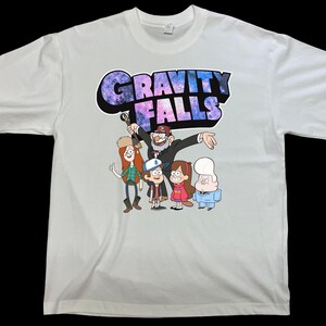 May include: White t-shirt featuring the text "GRAVITY FALLS" in a colorful, galaxy-themed font. The shirt displays a cartoon image of characters from the animated series. The design includes characters like Dipper, Mabel, and Grunkle Stan.