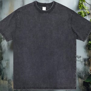 K&ouml;nnte beinhalten: Ein dunkelgraues T-Shirt mit kurzen &Auml;rmeln und Rundhalsausschnitt. Das Shirt hat ein verwaschenes, strukturiertes Aussehen. Der Hintergrund zeigt eine Backsteinmauer und Bl&auml;tter.