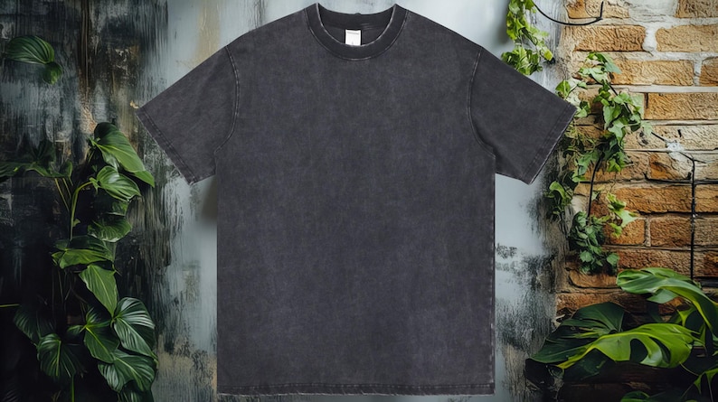 K&ouml;nnte beinhalten: Ein dunkelgraues T-Shirt mit kurzen &Auml;rmeln und Rundhalsausschnitt. Das Shirt hat ein verwaschenes, strukturiertes Aussehen. Der Hintergrund zeigt eine Backsteinmauer und Bl&auml;tter.