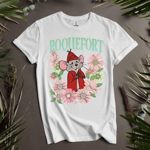 Peut inclure: T-shirt blanc avec un dessin de souris de dessin anim&eacute; portant un manteau et un chapeau rouges, entour&eacute;e de fleurs roses et jaunes. Le mot "ROQUEFORT" est imprim&eacute; au-dessus de la souris en vert clair.