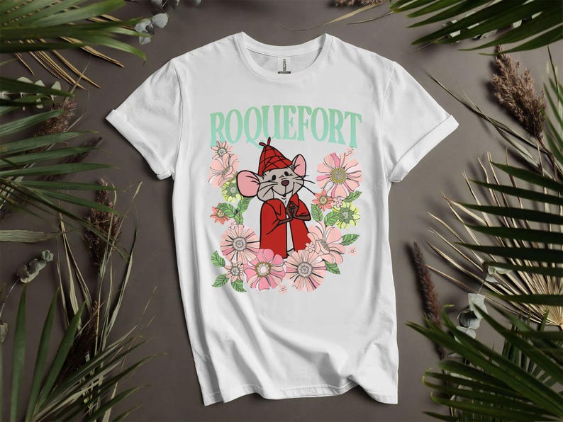 Peut inclure: T-shirt blanc avec un dessin de souris de dessin anim&eacute; portant un manteau et un chapeau rouges, entour&eacute;e de fleurs roses et jaunes. Le mot "ROQUEFORT" est imprim&eacute; au-dessus de la souris en vert clair.