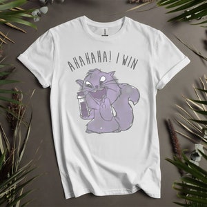 Puede incluir: Camiseta blanca con una ardilla de dibujos animados morada sosteniendo un vial y mostrando el dedo medio. El texto "AHAHAHA! I WIN" está impreso en gris encima de la ardilla. La camiseta se muestra sobre un fondo neutro con follaje verde.