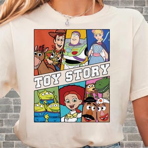 May include: Cream-colored t-shirt featuring a colorful graphic of characters from the animated film "Toy Story." The design includes Woody, Buzz Lightyear, Bo Peep, and other characters, with the words "TOY STORY" in bold letters.