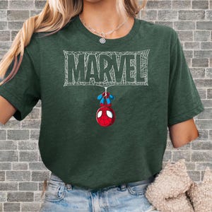 May include: Dark green t-shirt featuring the Marvel logo in white, with a Spider-Man graphic hanging below. The Spider-Man graphic is red, blue, and white. The shirt is a short-sleeved crew neck.