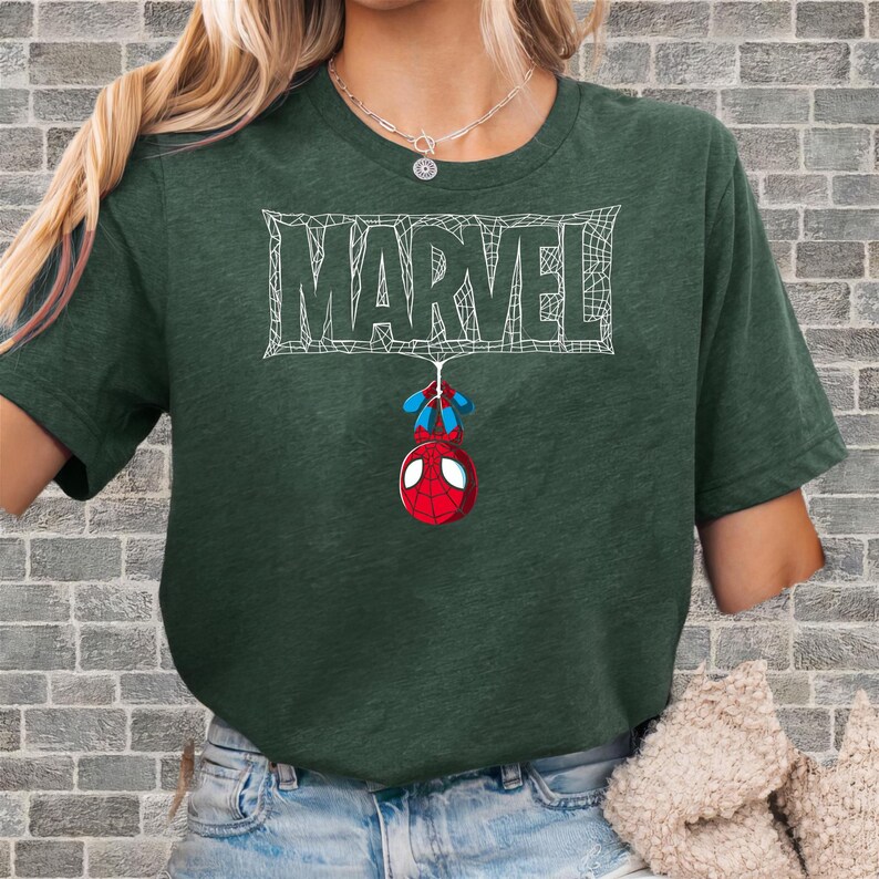 May include: Dark green t-shirt featuring the Marvel logo in white, with a Spider-Man graphic hanging below. The Spider-Man graphic is red, blue, and white. The shirt is a short-sleeved crew neck.