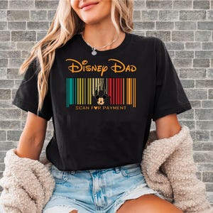 Peut inclure: T-shirt noir avec l'inscription "Disney Dad" en lettres dorées au-dessus d'un motif de code-barres coloré représentant Mickey Mouse et un château. Le texte "SCAN FOR PAYMENT" est en dessous.