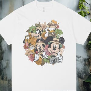 Puede incluir: Camiseta blanca con una ilustración colorida de Mickey Mouse, Minnie Mouse, Donald, Goofy y Pluto rodeados de flores y hojas. Mickey Mouse sostiene una cámara. El diseño es de estilo vintage.