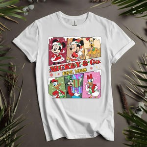 May include: White t-shirt featuring a colorful graphic of Mickey & Co. characters in Christmas attire. The design includes Mickey Mouse, Minnie Mouse, Donald Duck, Goofy, and Daisy Duck. The text "Mickey & Co. Est. 1928" is also displayed.