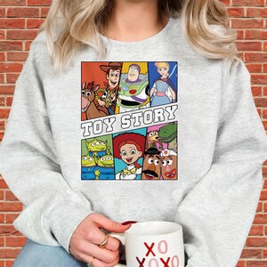 May include: A light gray sweatshirt featuring a colorful graphic of characters from the animated film "Toy Story." The graphic includes Woody, Buzz Lightyear, Jessie, and other characters, with the words "TOY STORY" in bold letters.