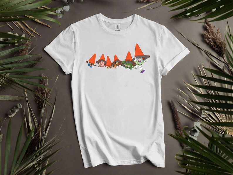 Puede incluir: Camiseta blanca con un dise&ntilde;o de dibujos animados de personajes que llevan sombreros de cono de tr&aacute;fico naranja. El dise&ntilde;o incluye a Buzz Lightyear y otras figuras en verde y marr&oacute;n. La camiseta es de material suave.