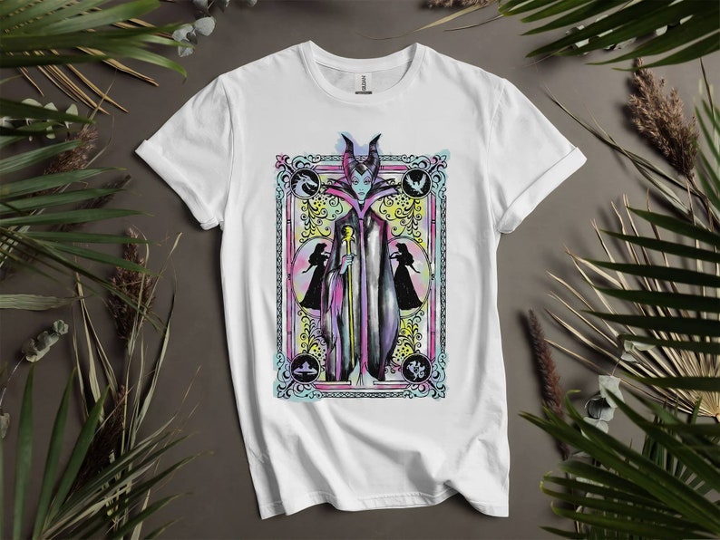 May include: White t-shirt with a colorful illustration of Maleficent from Disney's Sleeping Beauty. The illustration is framed in a decorative border with floral accents.