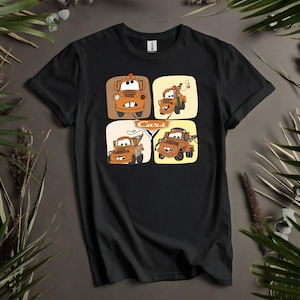 May include: Black t-shirt featuring a colorful graphic of four cartoon tow trucks in square frames. The word "Cars" is printed in white. The design includes orange, brown, and yellow elements, creating a playful and eye-catching design.