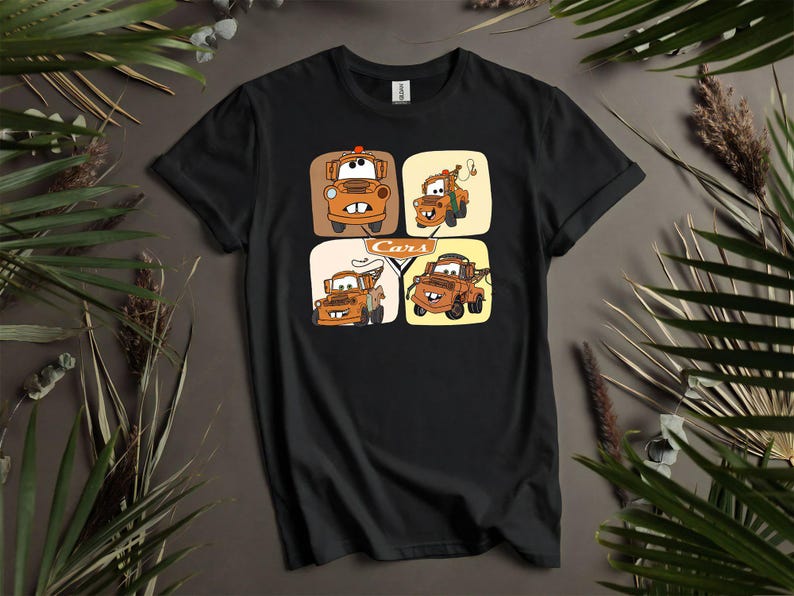 May include: Black t-shirt featuring a colorful graphic of four cartoon tow trucks in square frames. The word "Cars" is printed in white. The design includes orange, brown, and yellow elements, creating a playful and eye-catching design.