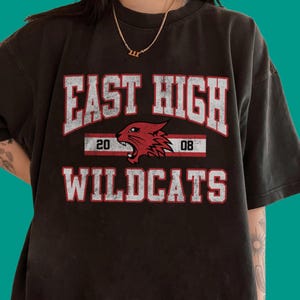 Op de afbeelding: Donkergrijs t-shirt met de tekst "EAST HIGH WILDCATS" in witte en rode letters. Het ontwerp bevat een rode wildcatkop en de cijfers "20" en "08". Het shirt heeft een vintage, versleten uitstraling.